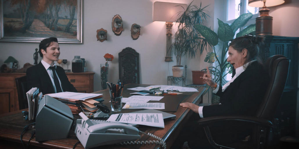 Wir sehen die beiden DarstellerInnen Gundi-Anna Schick & Frederic Stromenger in einer Kurzfilmproduktion in der Halbtotalen, wie sie sich an einem großen, hölzernen Schreibtisch gegenübersitzen.