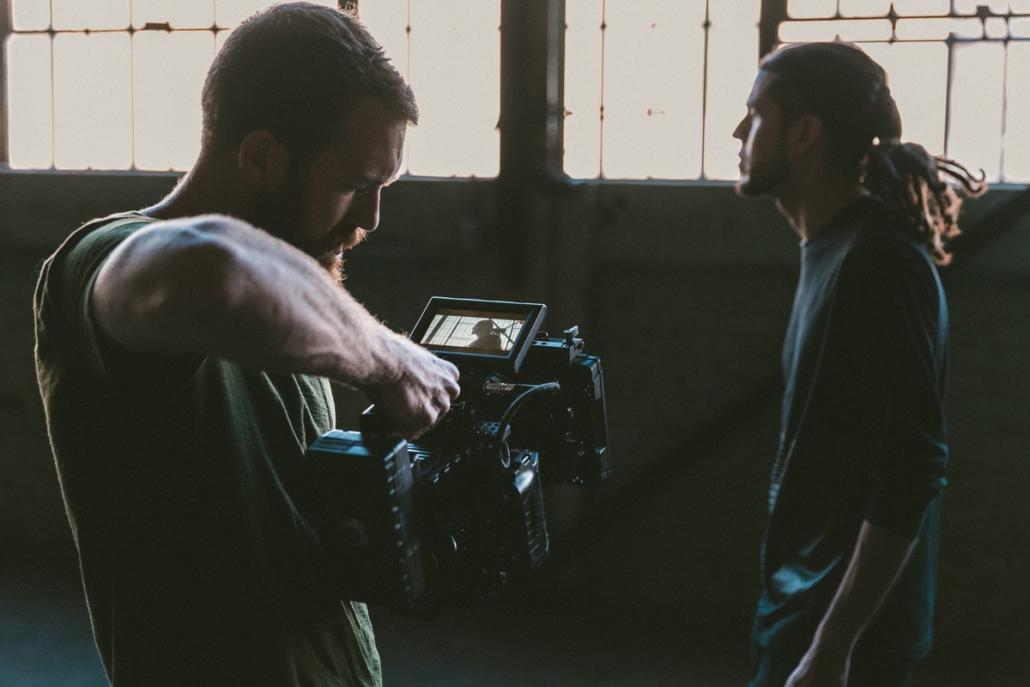 Ein Mitarbeiter vom Filmunternehmen Berlin hält per Hand eine Red-Kamera vom Bauch aus schräg nach oben, um einen mittelalten Protagonisten in der Halbnahen vor Fabrikfenstern zu zeigen