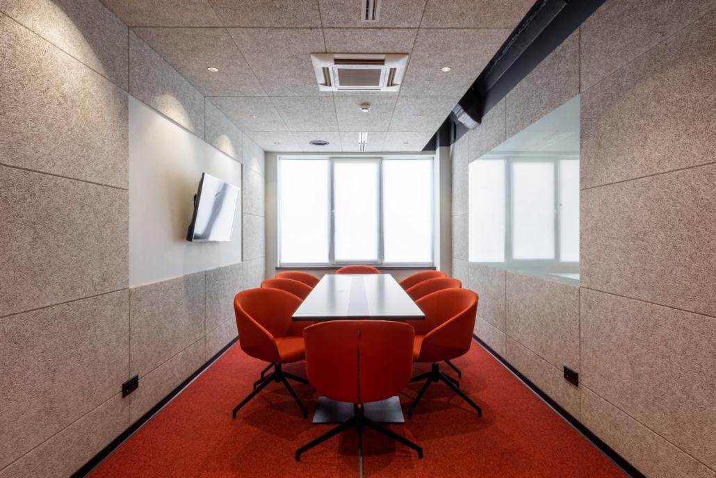 Einen Imagefilm wie in diesem Foto mit einem abgebildeten Meetingraum erstellen lassen. Wir sehen einen rechteckigen, modernen Meetingraum mit beigefarbenen, schalldichten Wänden, einem großen Flat-Screen an der linken Seite und großen Fenstern hinten an der Rückwand. Der Raum ist mit rotem Teppich ausgelegt und in der Mitte befindet sich ein großer, langer, rechteckiger Tisch, welcher von roten Stühlen in rundem Design umgeben ist. Das Bild zeigt den Raum mit Tisch und Stühlen in perfekter symmetrischer Anordnung.