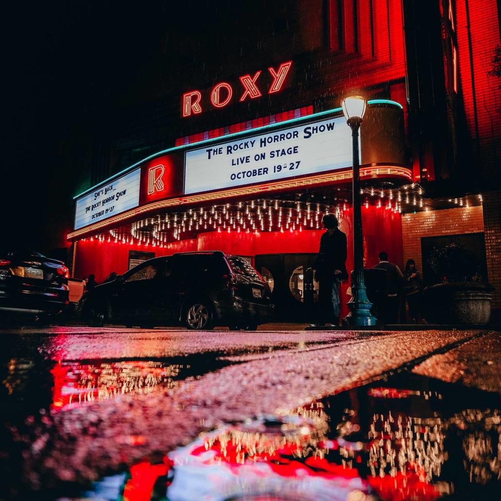 Möchte man die 10 besten Filme 2022 in besonders atemberaubender Atmosphäre genießen, so wäre dieses hier dargestellte Kino mit seiner prächtig ausgeleuchteten Außenfassade ein idealer Ort dafür: In Froschperspektive blicken wir von einer nassen Straße aus auf das mit zahlreichen Lichtern ausgestattete Kino. Auf dem Dach des Kinos ist in rot leuchtenden Buchstaben das Wort “Roxy” in Großbuchstaben als Name des Kinos zu sehen. Die vielen gelben und roten Lichter des Kinos spiegeln sich in den Pfützen im Vordergrund des Bildes wieder.