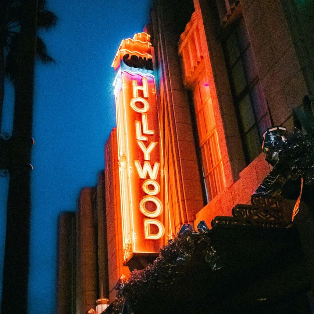 Abgebildet ist auf diesem Bild ein rot-orange strahlendes, großes Schild, welches die Aufschrift “Hollywood” in Großbuchstaben und in senkrechter Schreibweise zeigt und an einer Außenfassade erhöht angebracht ist. Es ist Abend, der Himmel dunkelblau und wir blicken auf das Schild schräg von unten vom Fußgängerbereich aus.