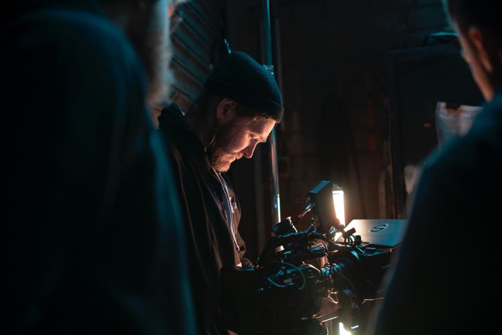 Für ein Werbevideo eine Filmproduktionsfirma aus Berlin beauftragen Dargestellt ist auf diesem Bild der Kameramann einer Filmproduktionsfirma Berlin in der Halbnahen, welcher gerade durch den Kontrollmonitor seiner Filmkamera blickt, um das aufgenommene Bild zu beurteilen. Links und rechts vorne im Bild sehen wir in der Unschärfe zwei weitere Crewmitglieder im Anschnitt.