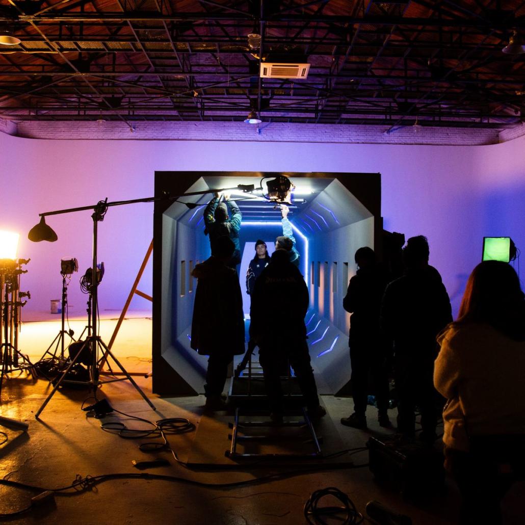 Gemeinsam mit einer Filmproduktionsfirma aus Berlin in einem Studio drehen In einem Studio können fiktive Filmformate sehr gut umgesetzt werden, wie auf diesem Foto verdeutlicht: Wir sehen die Crew einer Filmproduktionsfirma aus Berlin, welche gerade das passende Licht für den nächsten Take vorbereitet. Das Set ist magentafarben und bläulich ausgeleuchtet. In der Mitte des Sets befindet sich eine fiktive, futuristische Kulisse, vor welcher der Schauspieler steht, der von einer Kamera aufgenommen wird, die sich auf einem Dolly befindet.