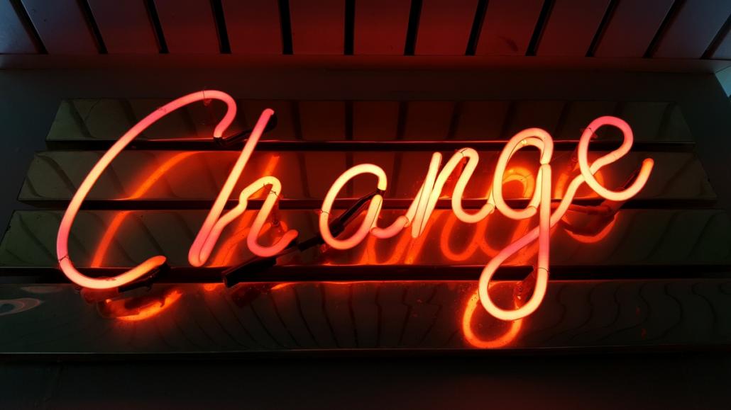 In Imagefilmen mit Neonlicht arbeiten, wie auf diesem Bild verdeutlicht: In einem dunklen Raum ist auf einer spiegelnden Oberfläche an einer Wand das in Schreibschrift geschriebene Wort “Change” zu lesen. Es besteht aus orange-gelb leuchtenden Neonröhren.