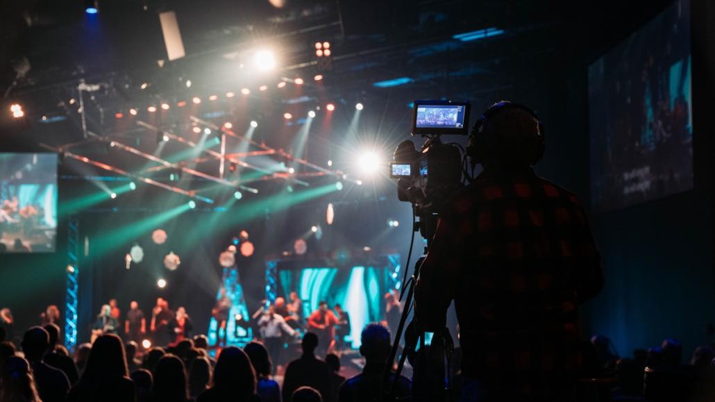 Von einem Livestream-Anbieter in Berlin eine Show live ausstrahlen Im Hintergrund ist eine große Bühne in der Unschärfe zu sehen, welche mit vielen grünen und roten Bühnen-Scheinwerfern ausgestattet ist, die sowohl Bühne als auch Publikum beleuchten. Das Publikum befindet sich hierbei im Bildmittelgrund. Recht vorne sehen wir, scharf abgebildet, im Bildvordergrund von hinten den Kameramann eines Livestream-Anbieters aus Berlin an seiner Studiokamera, welche sich auf einem Stativ befindet.