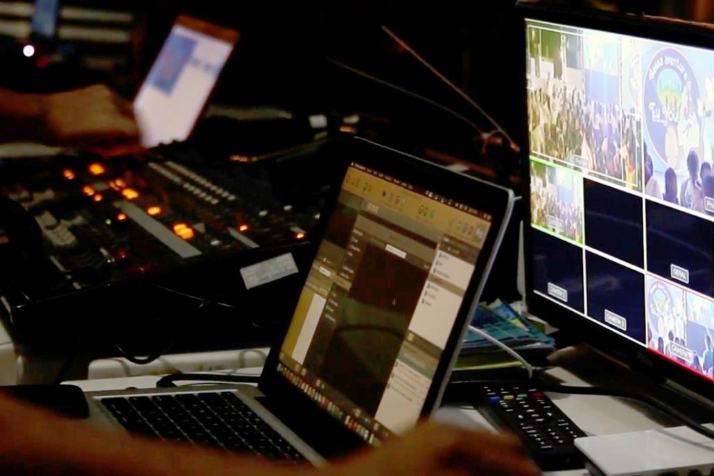 Für Shows und Konferenzen von einem Livestream-Anbieter in Berlin einen Livestream veranstalten Dargestellt ist auf diesem Bild der Regieplatz eines Livestream-Anbieters aus Berlin. Wir sehen rechts einen großen Programmbildschirm, mittig einen aufgeklappten Laptop mit laufender Streaming-Software, weiter links im Bild ein großes Mischpult in der Unschärfe und ganz hinten links im Bild einen weiteren aufgeklappten Laptop zusammen mit der Hand eines Mitarbeiters. Ebenfalls sehen wir links im Vordergrund unscharf die hand eines weiteren Mitarbeiters.