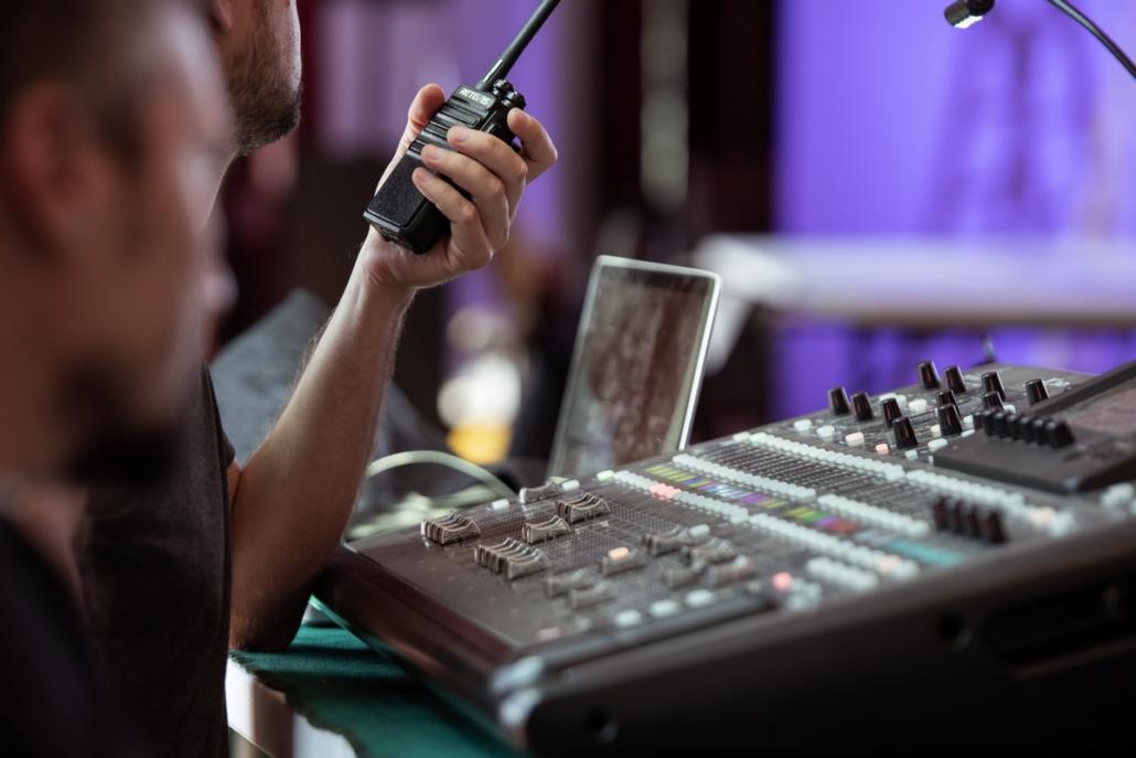 Als Livestream-Anbieter in Berlin eine Show live aufnehmen Im Fokus des Bildes sehen wir ein Tonmischpult und dahinter einen Laptop in der Unschärfe. Links im Anschnitt und in Nahaufnahme von der Seite gefilmt, sehen wir zwei Mitarbeiter einer Filmcrew. Der eine Mitarbeiter ist ebenfalls scharf abgebildet und hält ein Walkie-Talkie in seiner linken Hand hält. Der andere ist vorne links unscharf Over the Shoulder zu sehen.