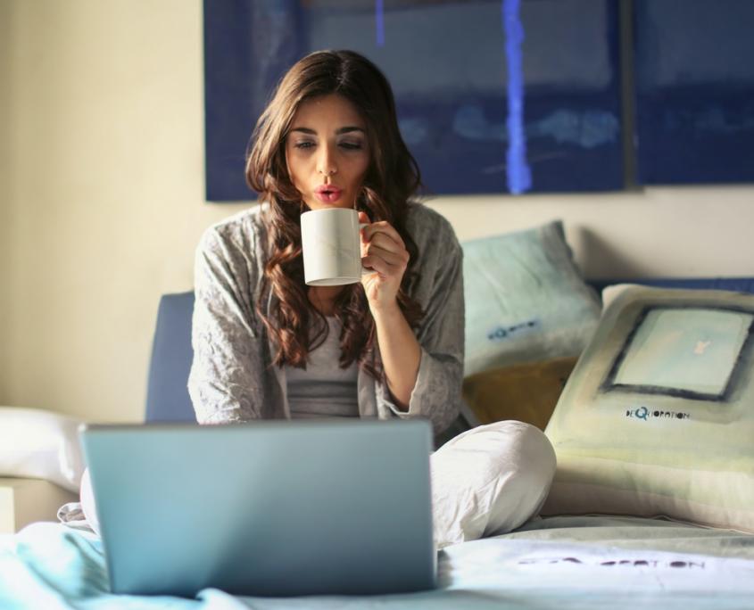 Eine junge Frau mit schwarzen Haaren sitzt im Schneidersitz im Bett und sieht sich Kaffee trinkend auf ihrem Laptop die besten Serien 2022 an.