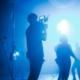 Auf dem Bild ist zentral der Kameramann einer Video-Produktionsfirma zu sehen. der an einem blau ausgeleuchteten Filmset mit tanzenden Darstellern im Hintergrund steht.