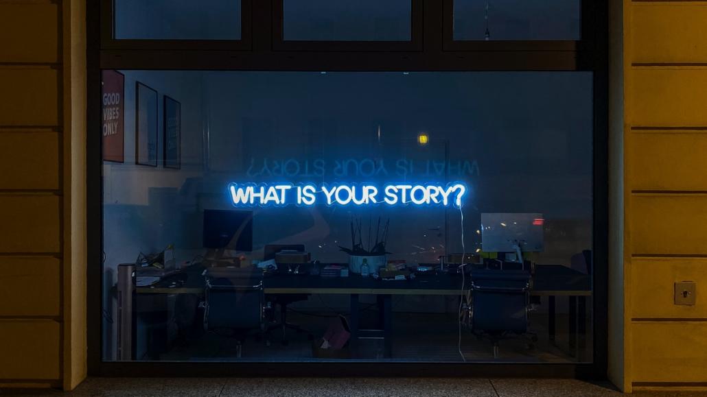 Ein blau leuchtendes Schild an der Fensterscheibe einer Erklärvideo-Agentur. An einer Fensterfront einer Erklärvideo-Agentur ist ein blau leuchtendes Schild angebracht, das aus dem Satz “What is your Story” in Großbuchstaben besteht.